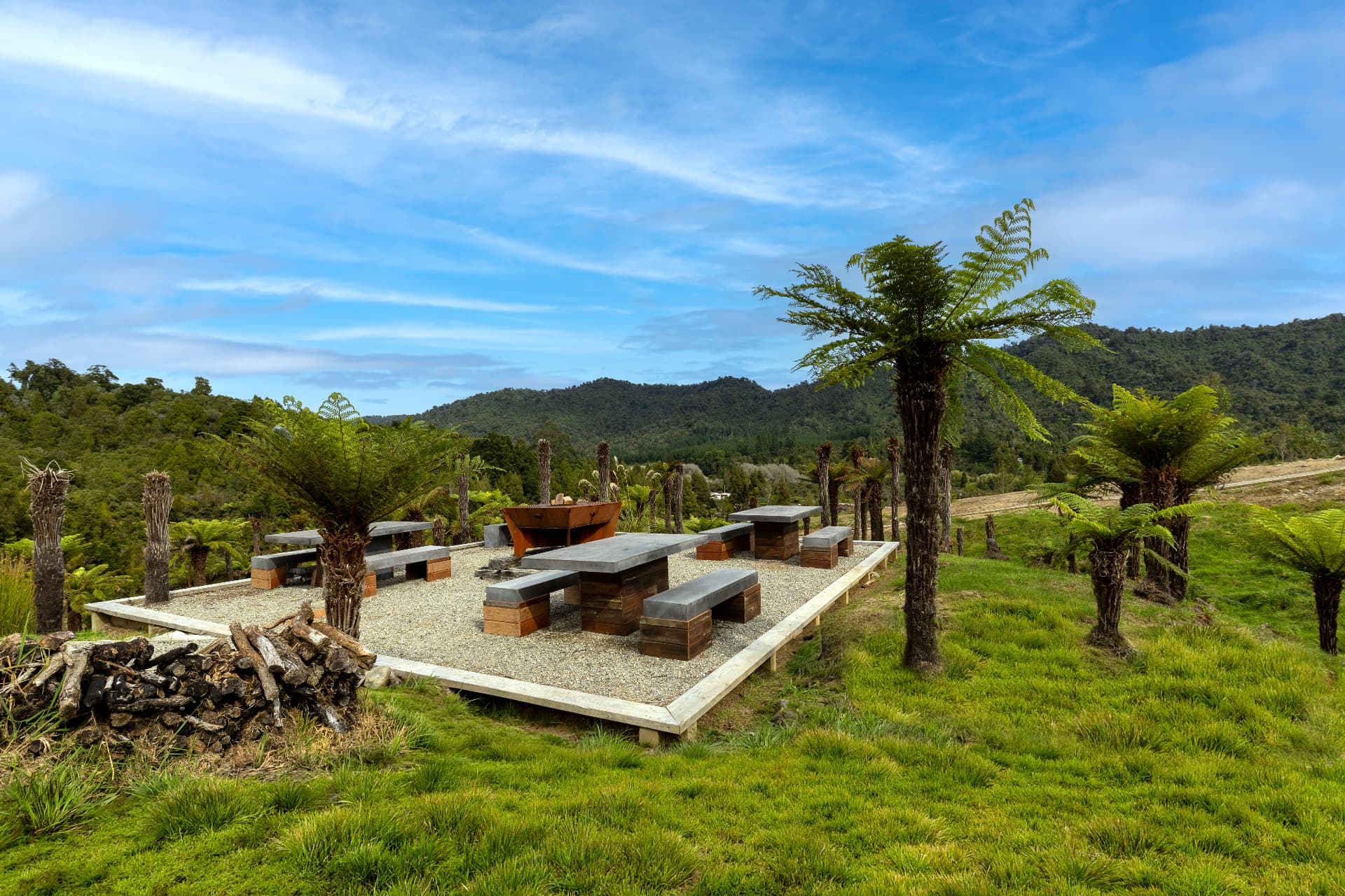 Outdoor dining terrace with concrete tables and a fire grill among tree ferns