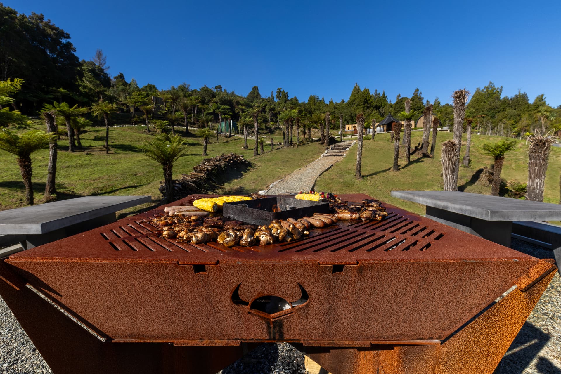 Corten steel grill loaded with corn, sausages and mushrooms above the village