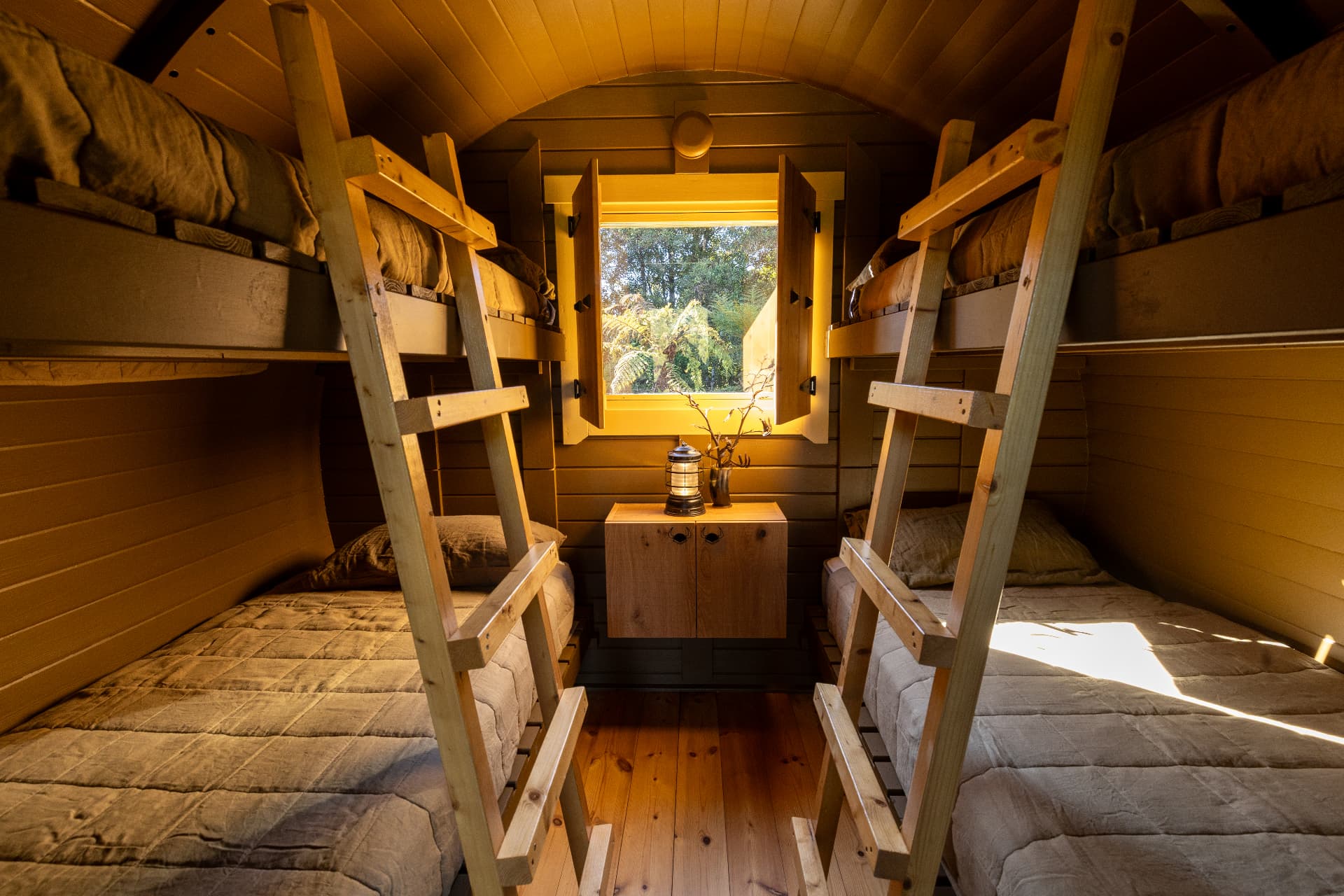 Interior of the Frigg barrel cabin with four timber bunks, ladders and a window framing tree ferns