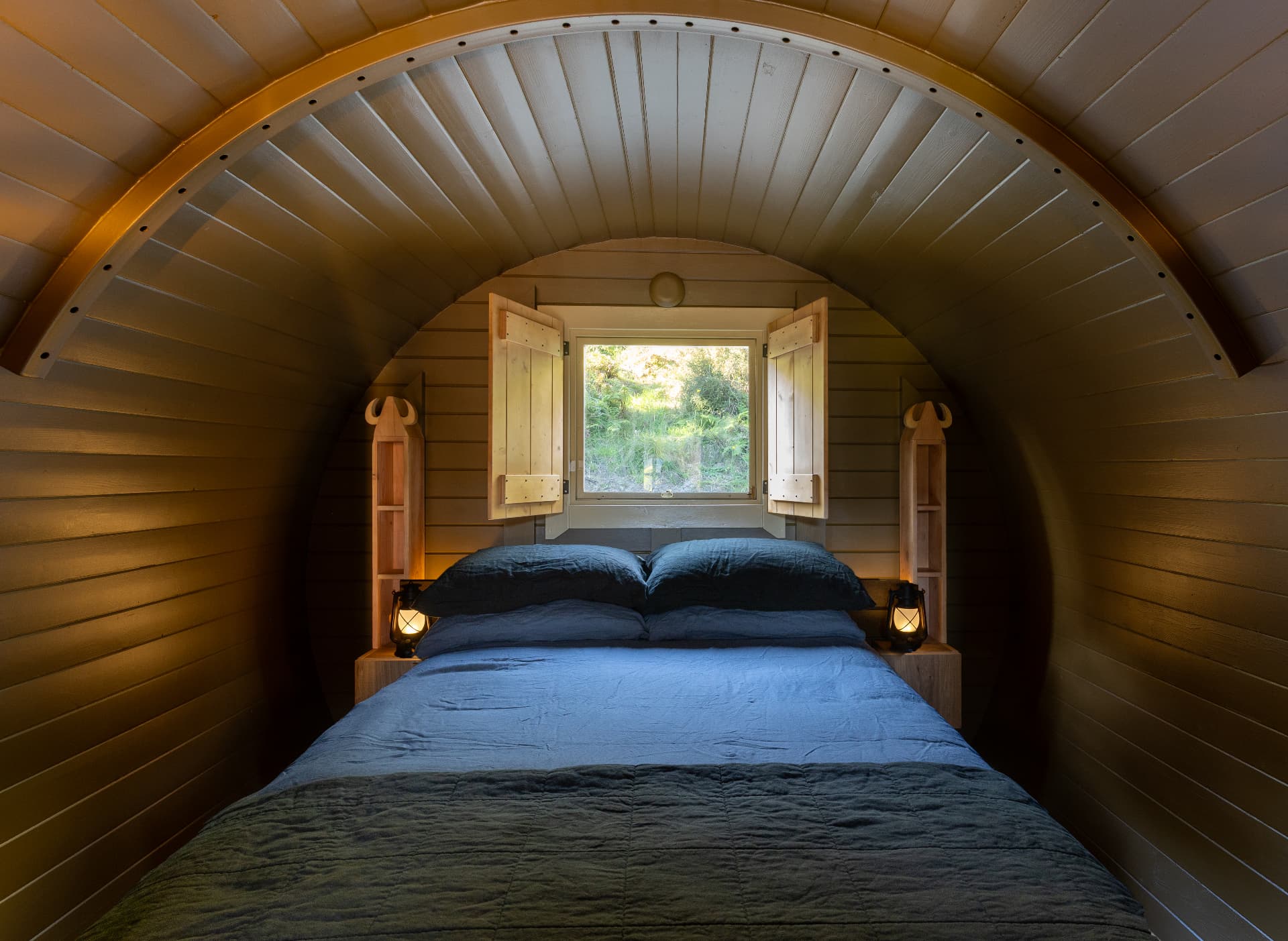 Interior of the Freyja barrel cabin with a double bed, lanterns and open shutters looking out to native bush